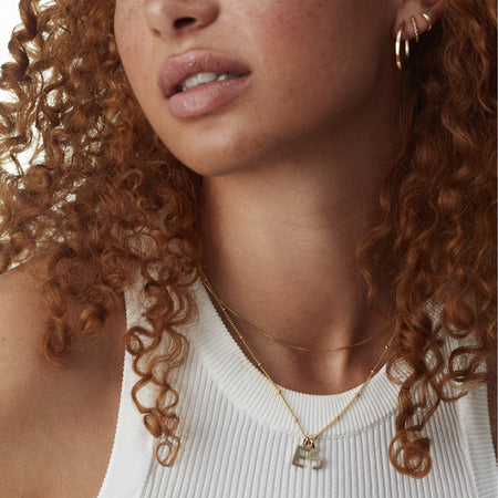 model wearing gold choker box chain layered with gold letter charms: 'E' and 'S' on a gold bead chain. model wears white vest
